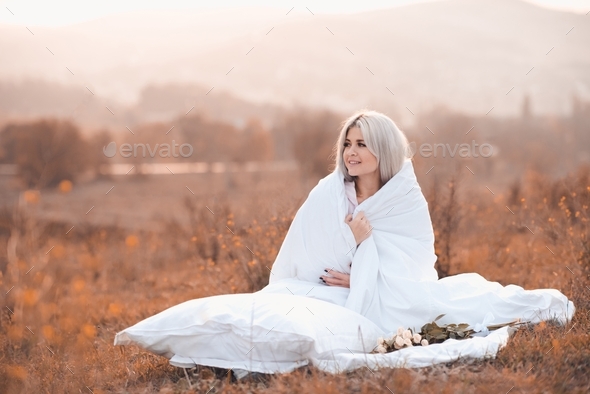 Happy young woman resting wrapped in white cozy duvet over autumn nature field outdoors. Fall season