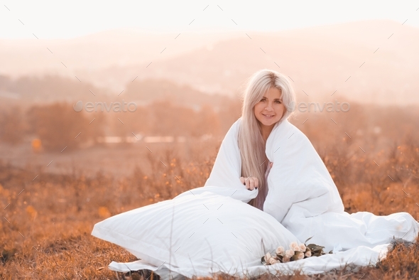 Happy young woman resting wrapped in white cozy duvet over autumn nature field outdoors. Fall season