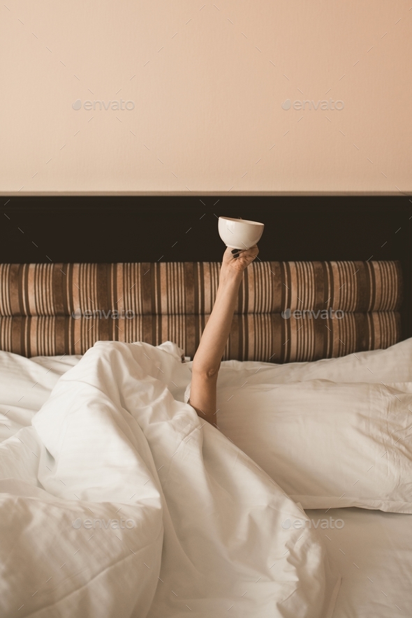 Girl lying in bed holding cup of coffee closeup in hotel room. Good sunday morning. Breakfast.