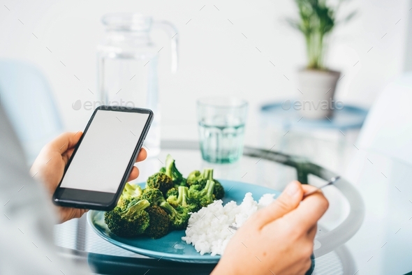 Woman Using Smart Phone While Eating Broccoli and rice at home.  - Stock Photo - Images