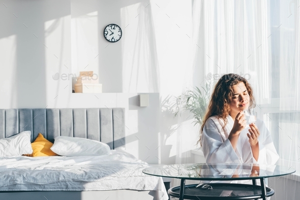 Pretty positive woman with long hair drinks water and sitting at glass table in spacious bedroom  - Stock Photo - Images