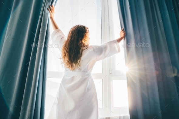 Woman opening curtains in room at sunrise. - Stock Photo - Images