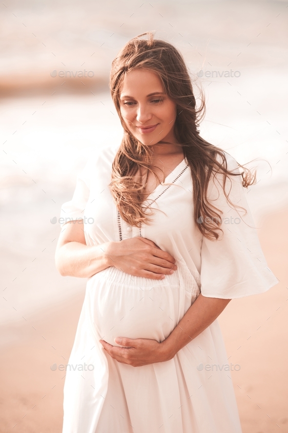 Blonde pregnant woman 24-26 year old wearing white dress holding tummy walking at beach outdoors.
