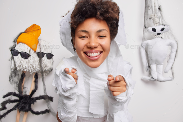 Positive Afro American woman pretends being ghost glad to celebrate halloween points at you smiles h