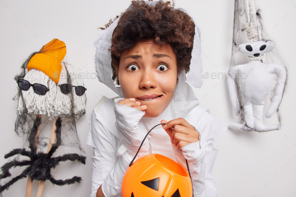 Scared worried curly haired African American woman holds carved pumpkin afraids of spooky creatures