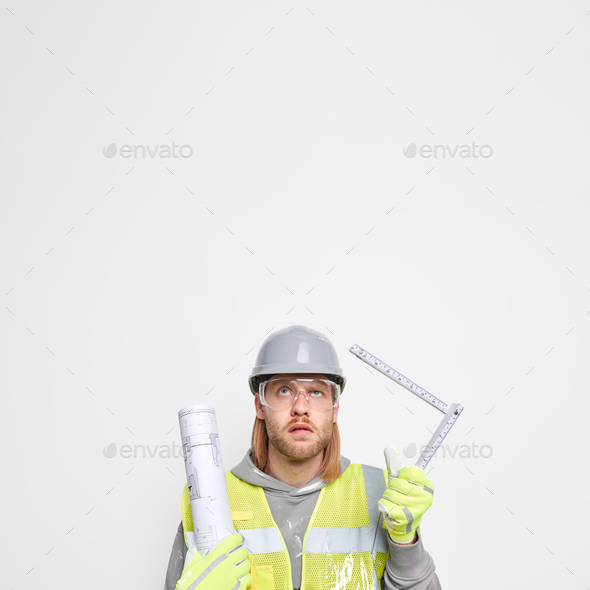 Vertical shot of ginger man maintenance worker holds tape measure and paper blueprint wears protecti