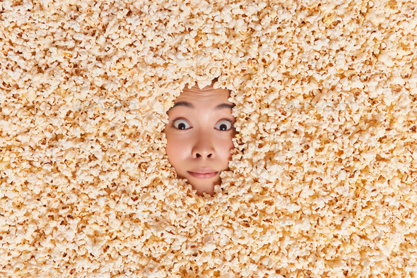 Womans face in popcorn. Surprised Asian woman showing only face covered with delicious sweet popped