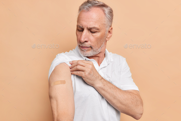 Old people protection and vaccination concept. Bearded European man looks at arm with plaster satisf