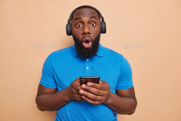 Shocked bearded man with dark skin holds modern gadget listens music via headphones downloads new so