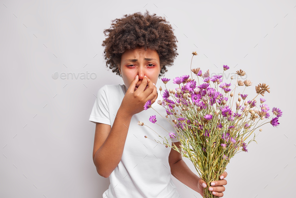 Curly haired sick young woman covers nose suffers from allergy symptoms holds bouquet of wildflowers
