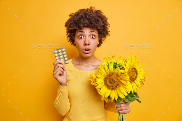 Startled curly haired woman holds pills and boquet of sunflowers being allergic to pollen has red sw