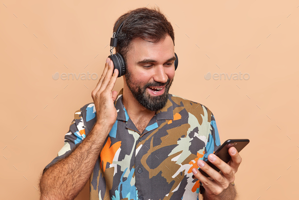 Pleased bearded man checks sound in stereo wireless headphones holds mobile phone downloads new song