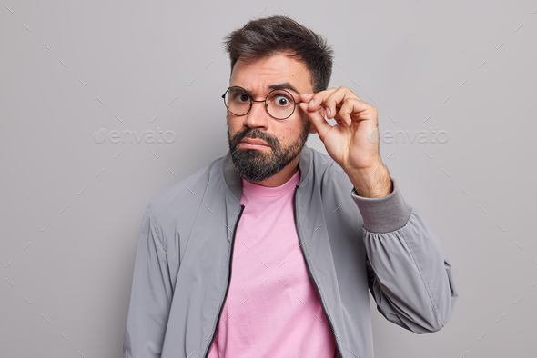 Serious bearded man keeps hand on rim of spectacles has attentive gaze directly at camera has surpri