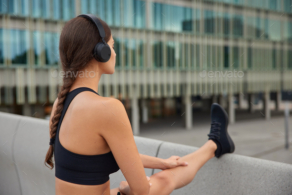 Back view of sporty woman in black cropped top sneakers stretches legs outdoors in modern environmen