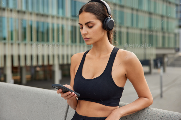 Waist up shot of sporty motivated female model downloads app for sport listens favorite music types