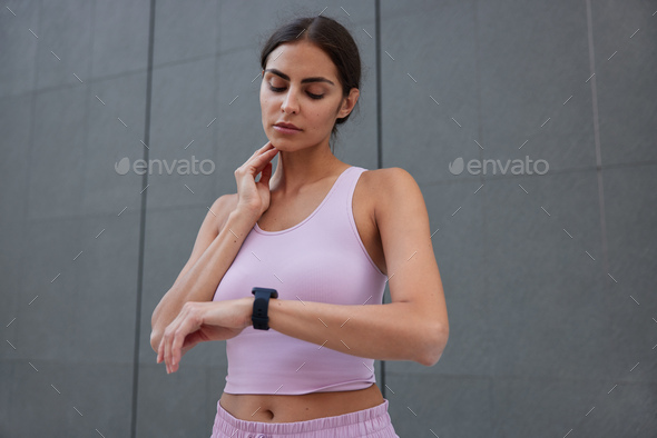Serious dark haired European woman controls pulse after training looks at smartwatch wears cropped t