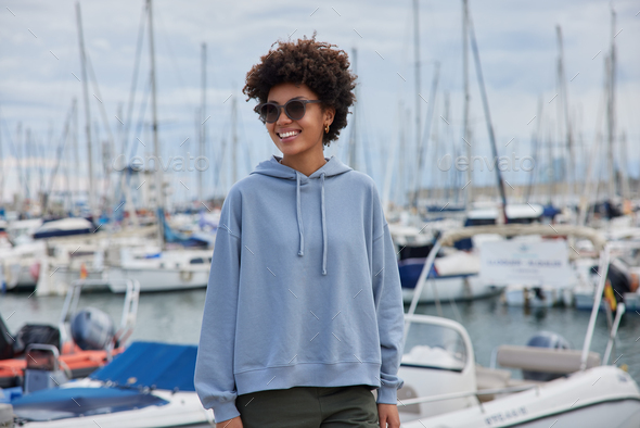 Happy Afro American woman with curly hair admires harbor during daylight wears trendy sunglasses hoo