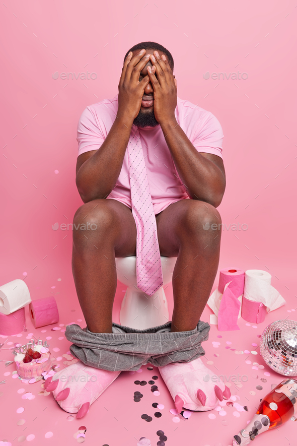 Dark skinned man poses comfortably on toilet bowl feels shy hides face with hands surrounded by part