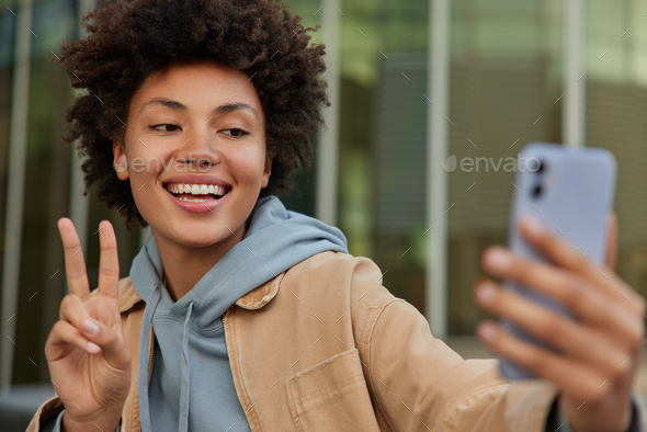 Carefree young positive woman makes peace gesture takes selfie on smartphone camera makes online cal