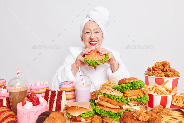 Pleased elderly woman in white bathrobe and towel wrapped on head eats delicious burger has cheat me