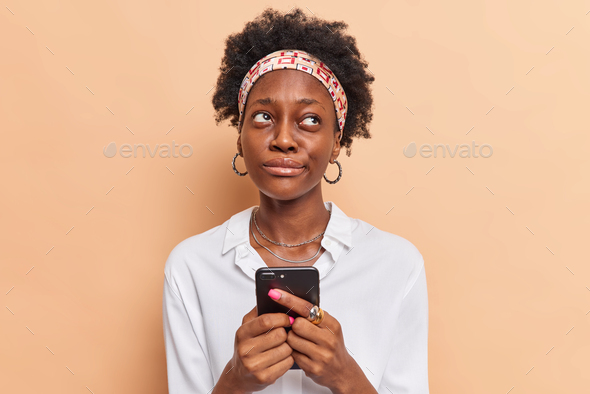Thoughtful dark skinned woman holds mobile phone ponders how to answer question under post on websit