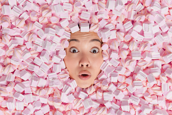 Stunned Asian woman stucks head through pink and white marshmallow stares bugged eyes at camera find