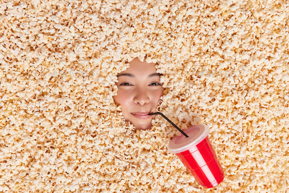 Satisfied Asian woman eats popcorn and drinks cola enjoys free time has lazy day cosumes tasty favor