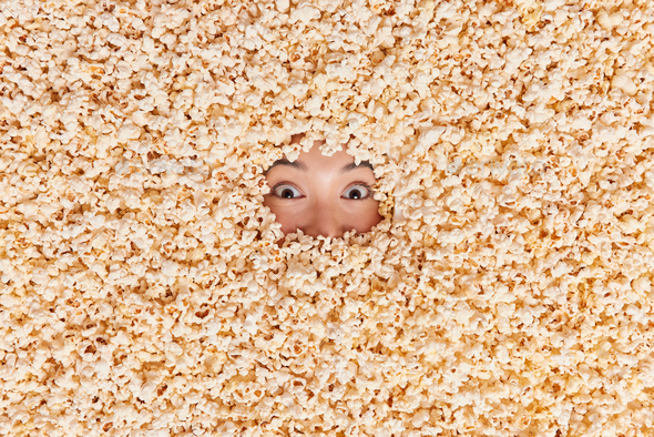 Surprised female model looks out of popcorn. Woman buried in air popped corn shown only eyes has sho