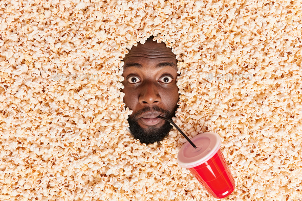 Startled black bearded man drowned in popcorn drinks soda watches film in cinema has surprised expre