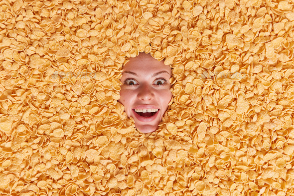 Overhead shot of surprised cheerful young woman sticks head through dry cornflakes keeps mouth opene