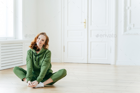 Photo of delighted redhead female sits on lotus pose, wears tracksuit, being in good body shape