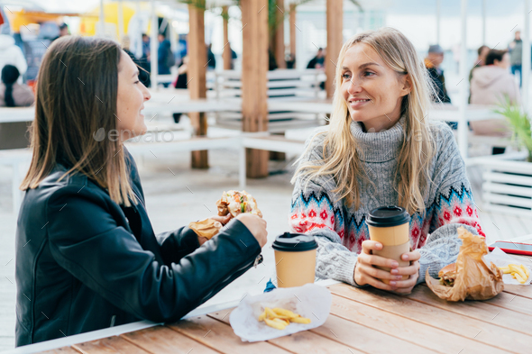 Two female friends eat hamburgers, drink coffee and chat on the street, sitting at the table.