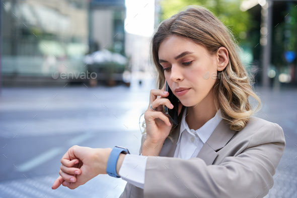 Serious businesswoman checking time on watch and calling person on mobile  phone, waiting for Stock Photo by benzoix