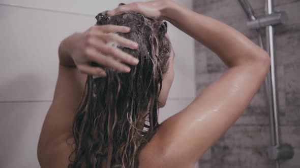 Back Look of Woman Washes Head with Shampoo Under Shower alt