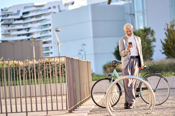 Old senior man using phone using bike rental app renting bicycle in city park.