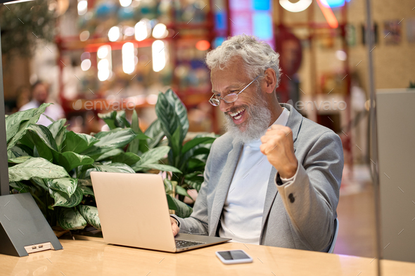 Happy older business man using laptop celebrating good results winning online.