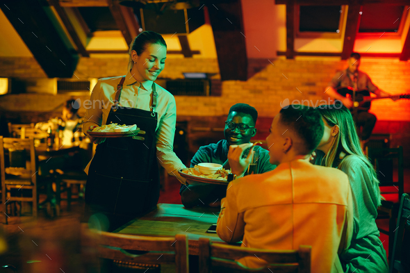 Young happy waitress serving snack to group of friends in a pub.