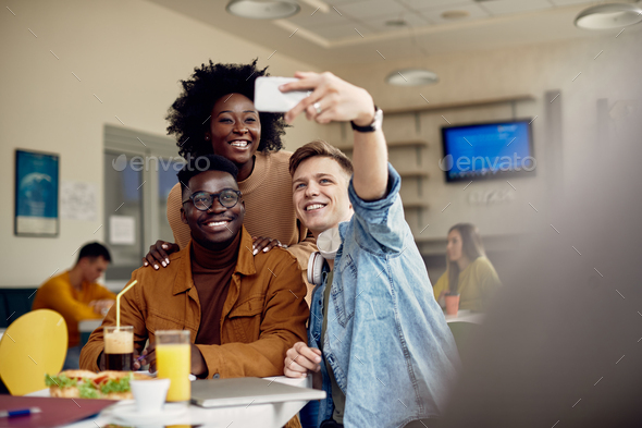 Happy multi-ethnic group of students taking selfie on lunch break in cafeteria.