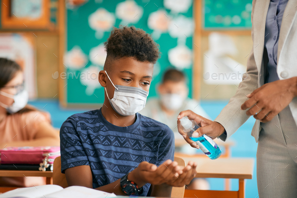 Black schoolboy wearing face mask and disinfecting his hands in classroom due to COVID-19 pandemic.