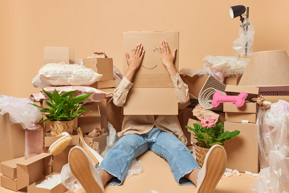 Unrecognizable woman covers head with cardboard box poses on floor surrounded by personal belongings