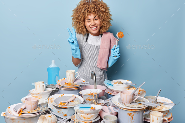 Happy curly housewife wears apron washes up dishes with brush poses near sink full of dirty plates