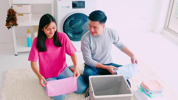 Husband helping his wife folding clothes. Happy Asian couple spending time together at home. alt