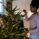 Smiling african female stringing LED garland along branches of live Christmas tree at cozy home - PhotoDune Item for Sale