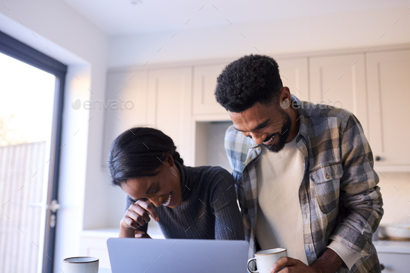 Laughing Young Couple At Home Looking At Laptop On Counter In Kitchen Together - Stock Photo - Images