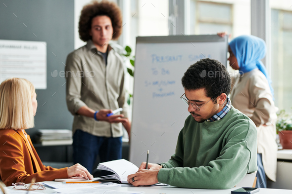 Young man making notes in copybook while sitting by desk at lesson