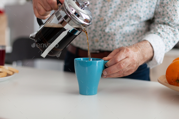 Retired man holding french press pouring hot coffee in cup during breakfast