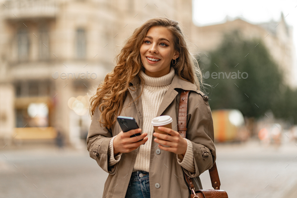 Young woman with curly blonde hair using the phone with a cup of coffee in hands on the city streets - Stock Photo - Images