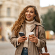 Young woman with curly blonde hair using the phone with a cup of coffee in hands on the city streets - PhotoDune Item for Sale