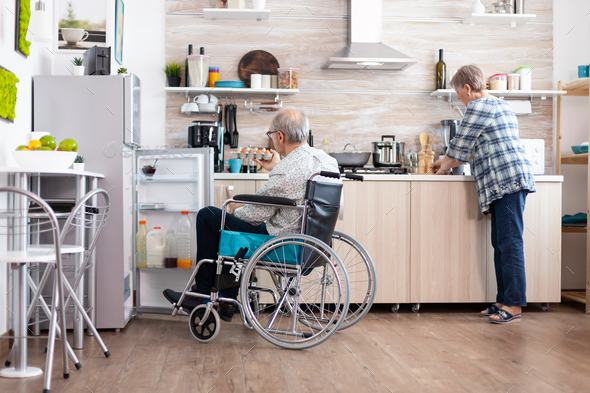 Handicapped man in wheelchair opening refrigerator and helping wife preparing breakfast