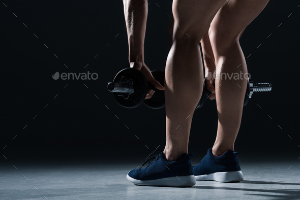 low section view of female bodybuilder in sneakers training with dumbbells, on black